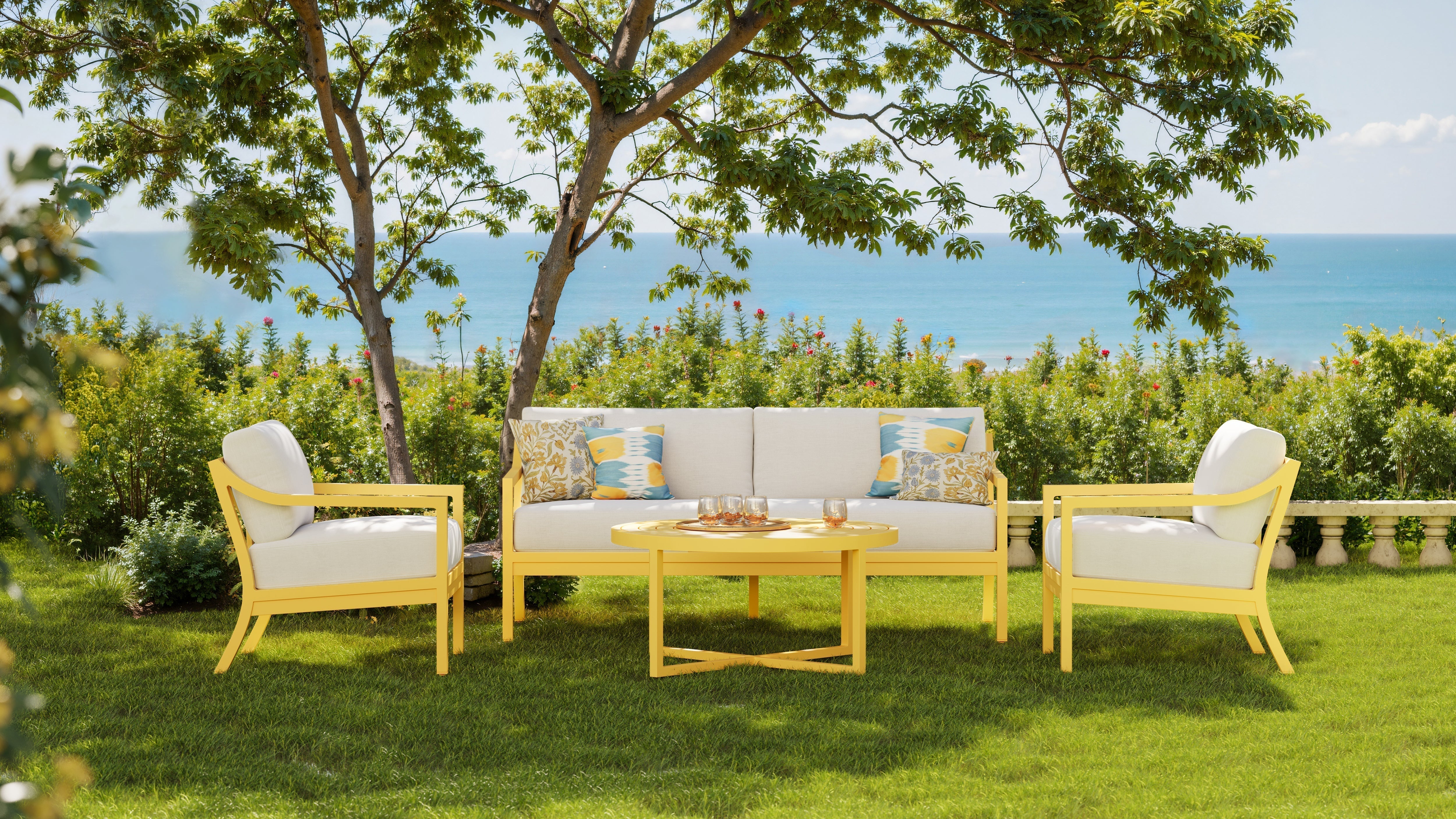 A coastal outdoor seating area featuring a yellow-framed sofa and armchairs with white cushions, accented by patterned throw pillows and a round yellow coffee table, set under a leafy tree with ocean views in the background.