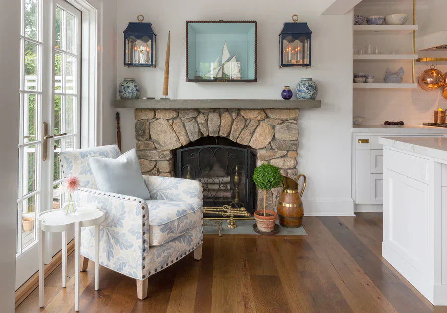 A small white Tini Table in a cozy living area with a stone fireplace, and light blue patterned armchair.